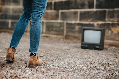 Free Back view of anonymous female in jeans standing on ground against stone wall and old fashioned television on blurred background Stock Photo