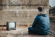 一个穿着外套、面目全非的男性背靠涂鸦的破旧混凝土墙坐在地上，一边看着背景模糊的老式电视屏幕