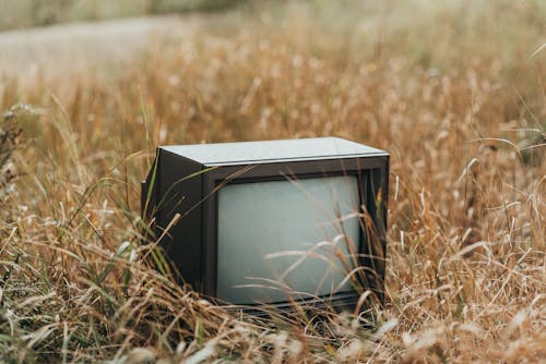 Small black obsolete old fashioned television placed on withered dry grass on street against blurred background in countryside in nature