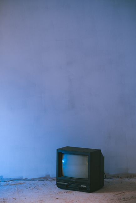 Small black old fashioned television placed on concrete floor ground near shabby white wall at dim blue light in evening time