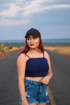 A young woman stands confidently on a deserted road in casual summer attire, expressing bold style.