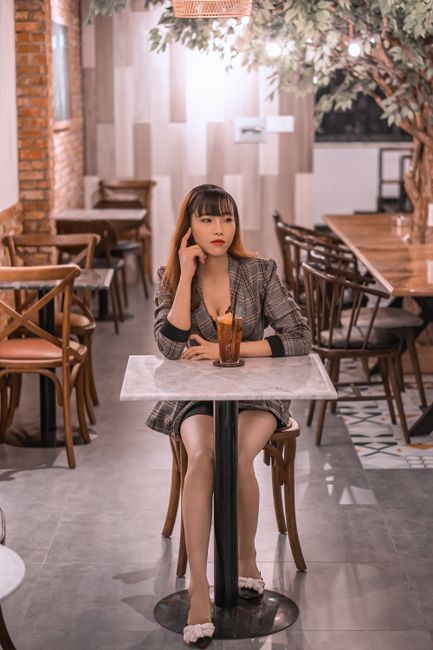 Young pondering ethnic female in elegant apparel touching face at table with refreshing drink while