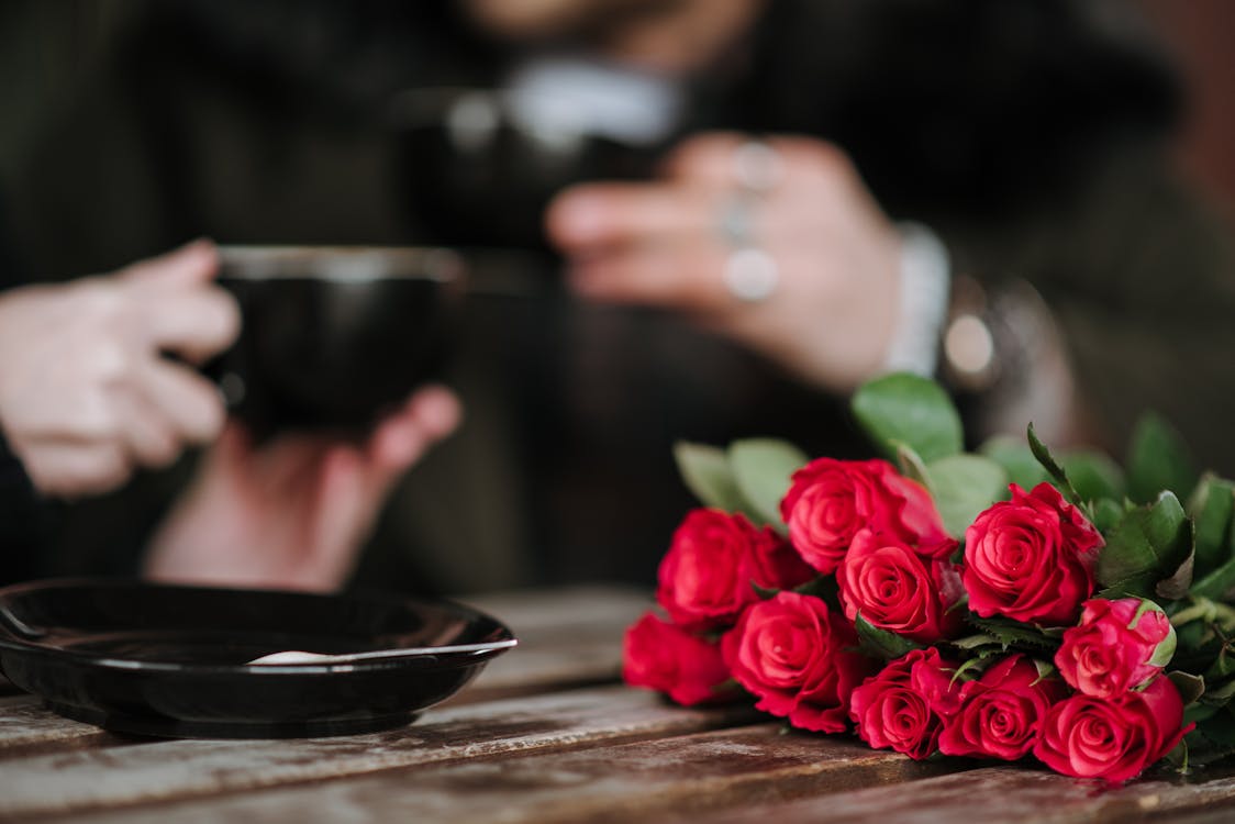 Crop unrecognizable couple with cups of beverages at wooden table with blossoming flowers in cafeteria