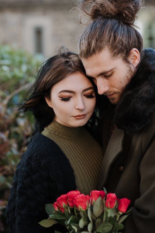 Free Hipster man with hair bun embracing charming girlfriend with closed eyes and blooming flowers in city Stock Photo