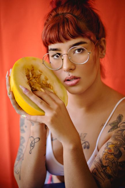 Portrait of a tattooed woman with red hair holding a melon, set against a bold red background.