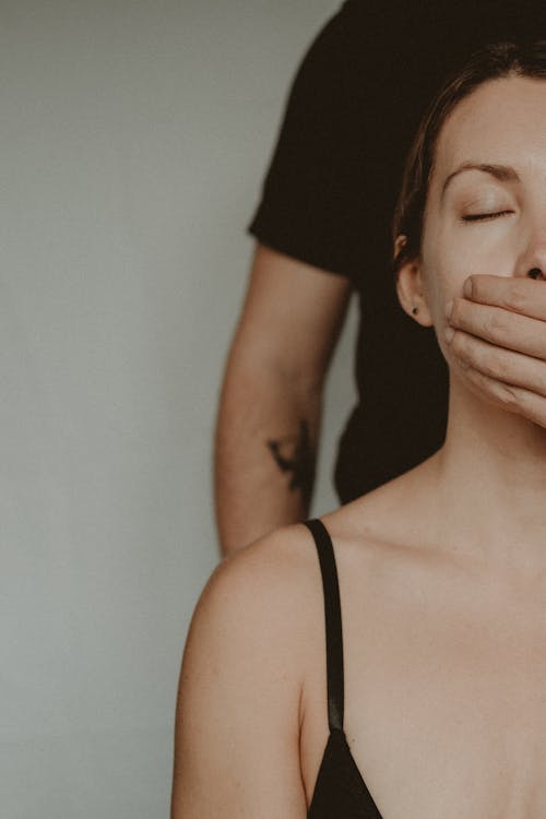 Crop abusive man standing behind back and closing mouth with hand of female with closed eyes while standing on white background