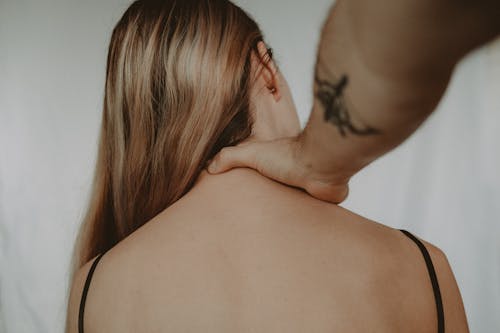 Anonymous abusive man with tattoo touching neck of vulnerable woman sitting on white background during domestic violence in light room