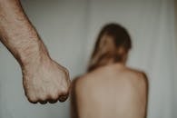 Aggressive abusive anonymous man clenching fist while insulting unrecognizable vulnerable female sitting on blurred background in light room during violence