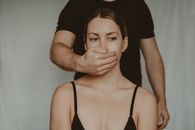 Unrecognizable male covering mouth of frighted vulnerable female with hand while standing behind back on white background in light room