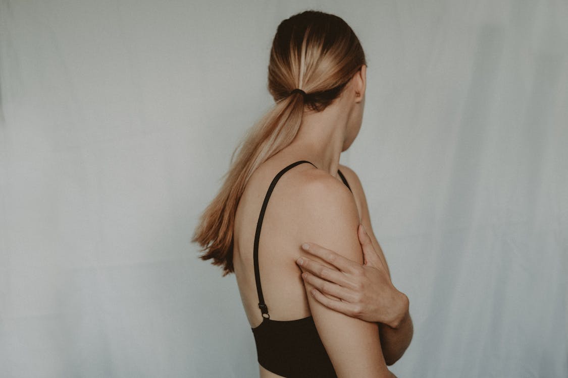 Side view of unrecognizable young female with long hair and black bra standing near grey wall turning head away