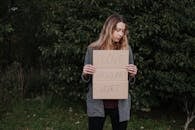 Lonely upset female in casual outfit standing near trees and grass while holding cardboard against abuse with inscription Love Should Not Hurt