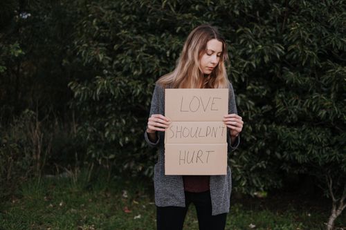 Free Lonely upset female in casual outfit standing near trees and grass while holding cardboard against abuse with inscription Love Should Not Hurt Stock Photo