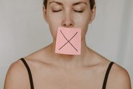 Young woman in black lingerie with closed eyes showing pink sticker with black cross against violence and abuse on lips on white background in light studio