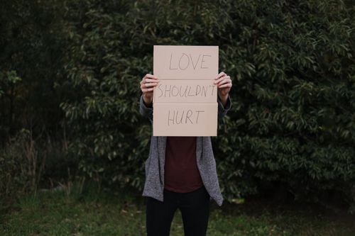 Free Faceless lady in casual outfit standing near green bushes and showing cardboard saying Love Should Not Hurt Stock Photo