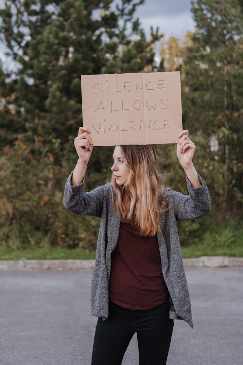 Woman in casual clothes standing on road near green trees and demonstrating cardboard poster saying Silence Allows Violence