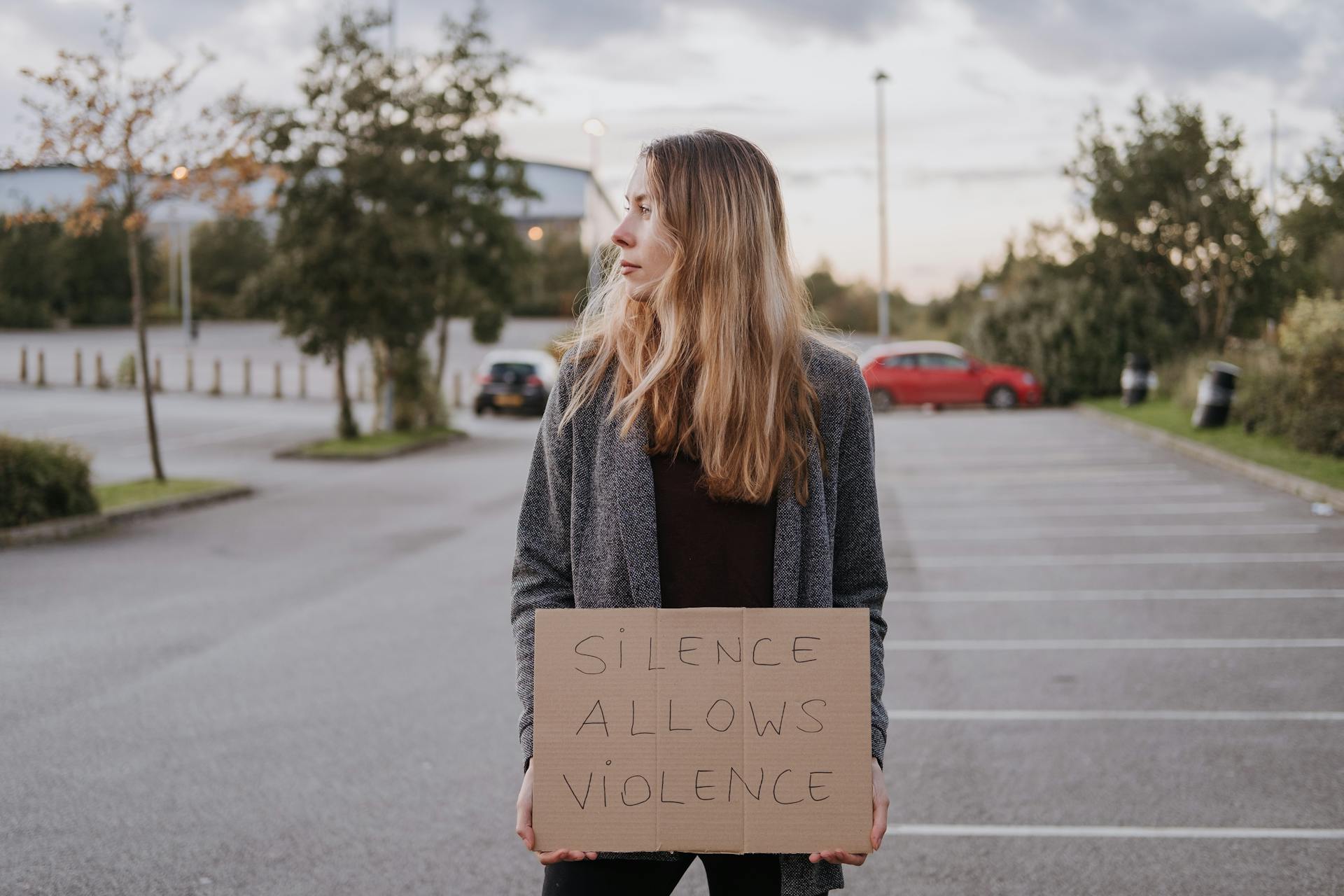 Young woman outdoors holding a protest sign with 'Silence Allows Violence' in a parking lot.