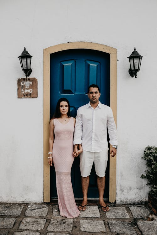 Full body of confident young ethnic couple in stylish clothes holding hand and looking at camera while standing near blue door of cozy white house