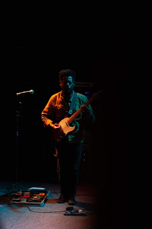 Full body of talented musician playing electric guitar on stage near microphone stand while performing song during live concert in darkness