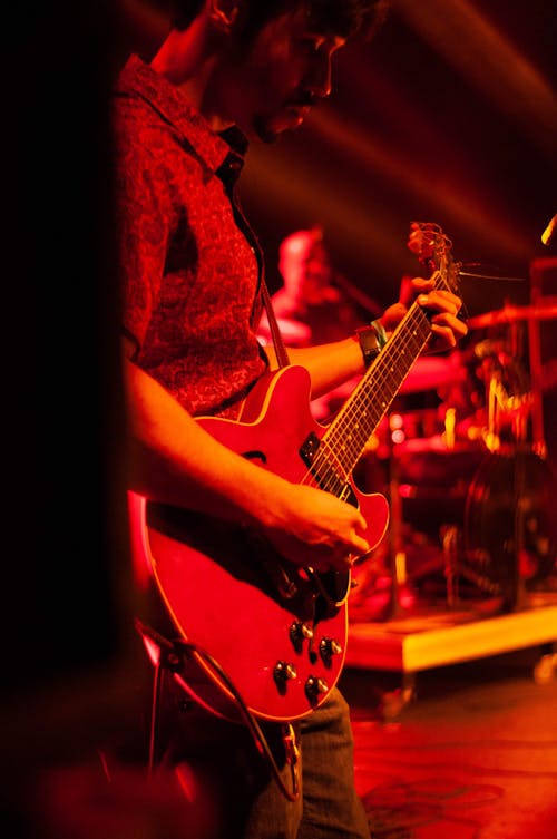 Free Side view of crop male musician playing electric guitar while standing on stage with drummer on blurred background and red illumination during concert Stock Photo
