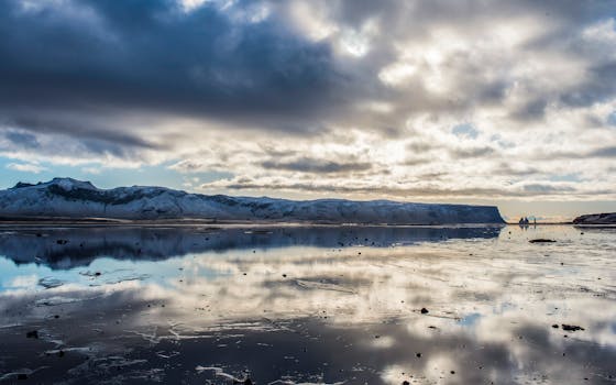Stunning Icelandic landscape with snow-covered mountains reflected in a tranquil ocean under a dramatic sky.