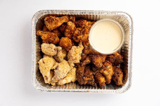 Delicious assorted fried chicken nuggets with dipping sauce in a foil container.