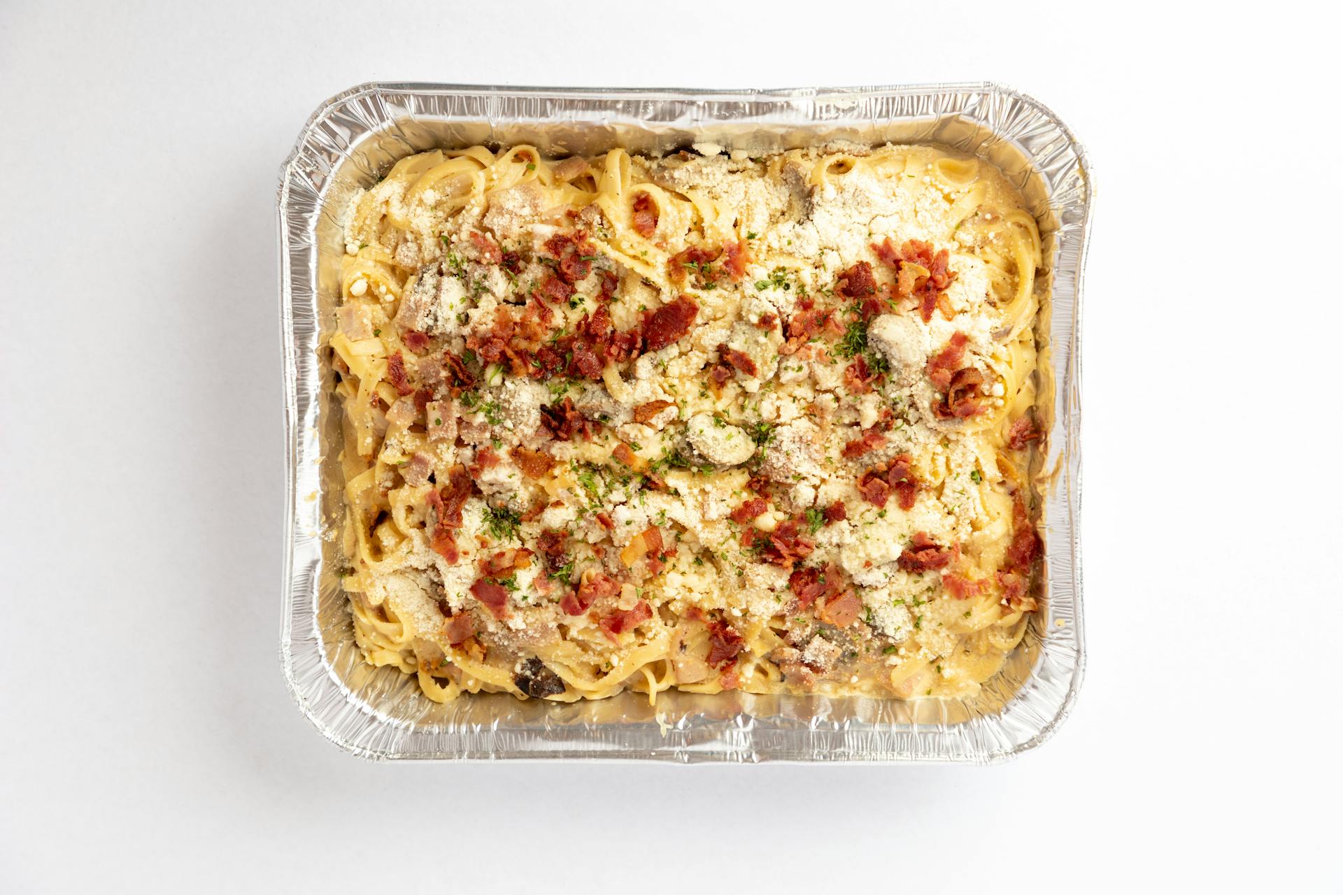 Pasta carbonara with bacon and cheese in an aluminum tray on a white background.