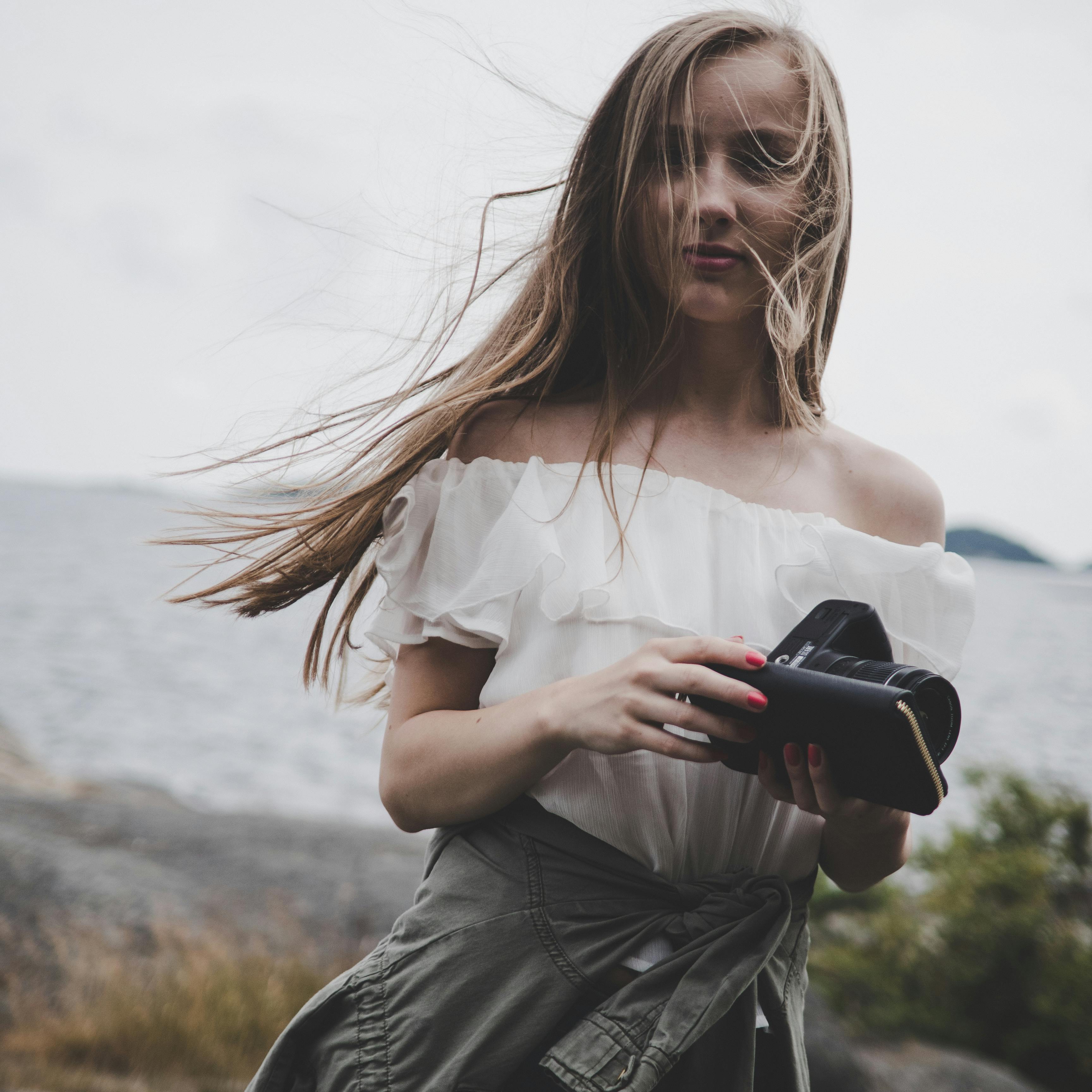A stylish young woman holding a camera by the seaside, embracing the breeze.