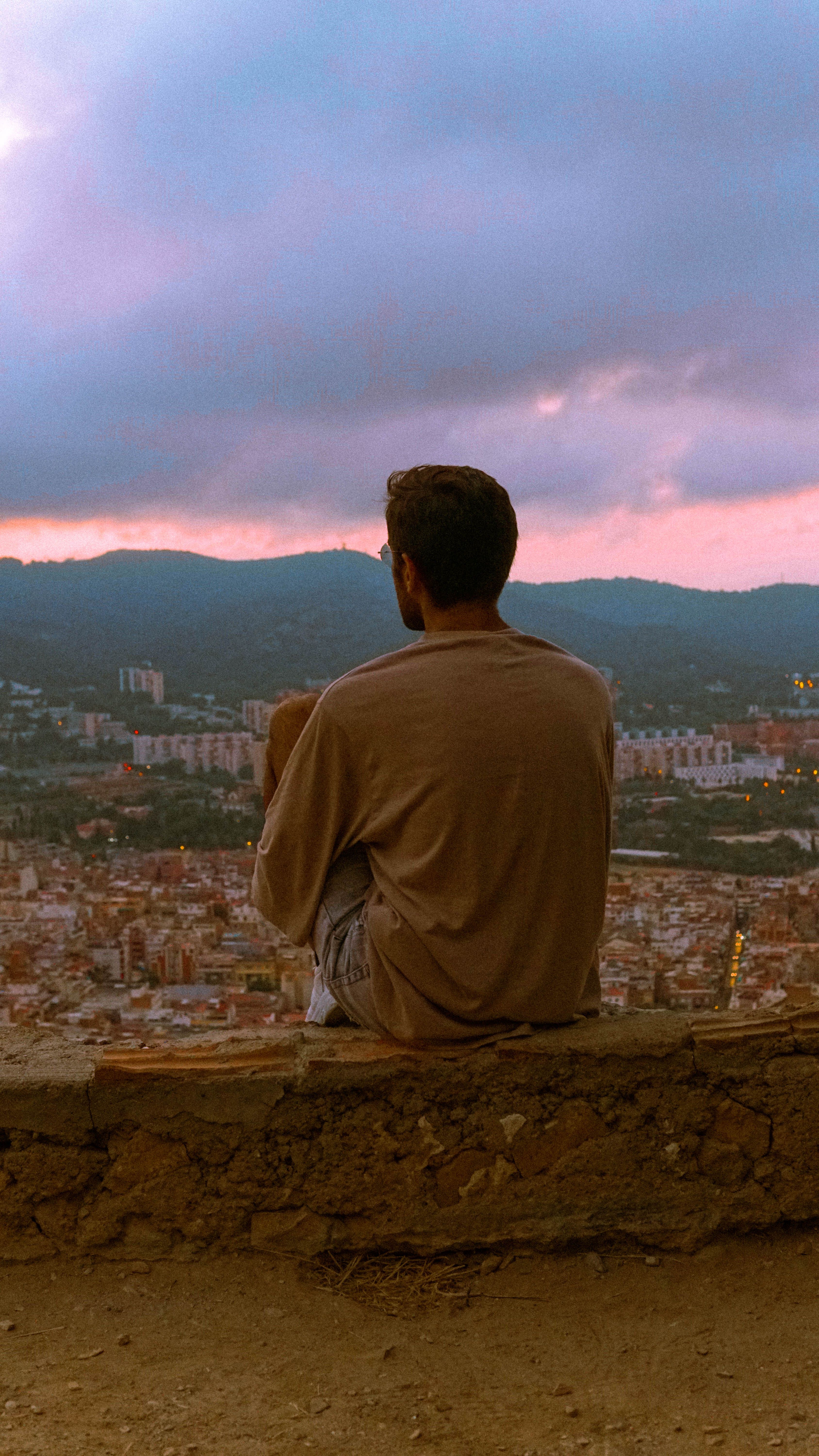 Back view of a man sitting on a rock with a dramatic sunset over the cityscape.