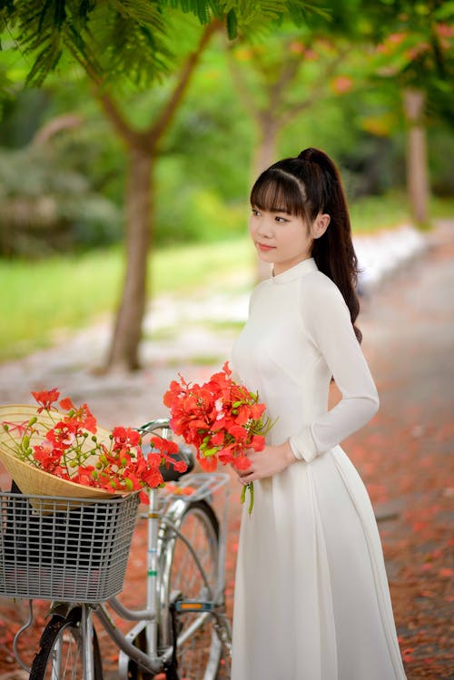 Woman in White Dress Holding Flowers Standing Near the Bicycle
