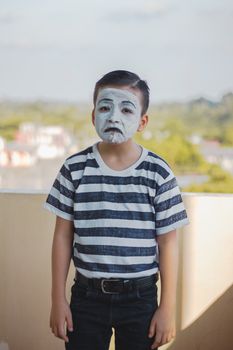 一个脸上涂着油漆的小男孩，穿着条纹衬衫在户外，看起来很悲伤