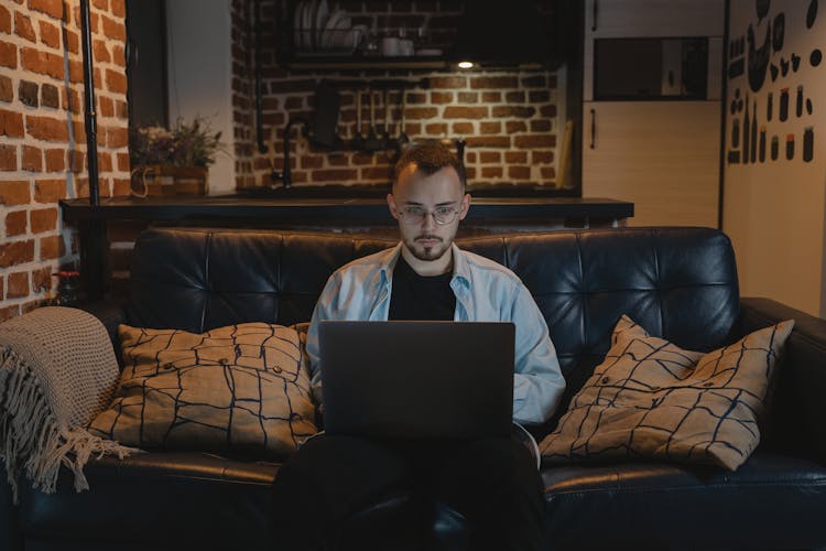 Man In Black Crew Neck T-shirt And Denim Jacket Sitting On A Couch Using His Laptop 