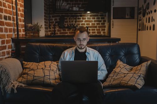 Free Man working remotely on a laptop in a cozy, stylish apartment with exposed brick walls and modern decor. Stock Photo