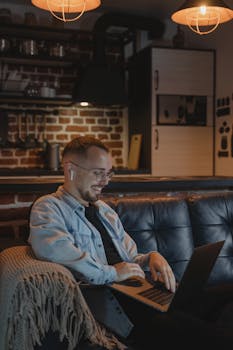 A young man smiling while working remotely on a laptop at night in a cozy indoor setting.