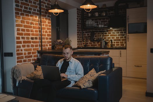 A bearded man sits comfortably on a couch, working on his laptop in a stylish living room.