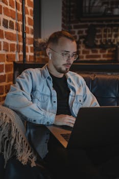 Man in denim jacket using a laptop at home with a cozy brick wall backdrop.