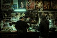 Two musicians focusing on guitar repairs in a Santiago workshop, surrounded by posters and tools.