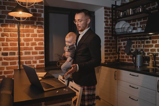 A father multitasks with a baby while working on his laptop in a cozy kitchen setting.