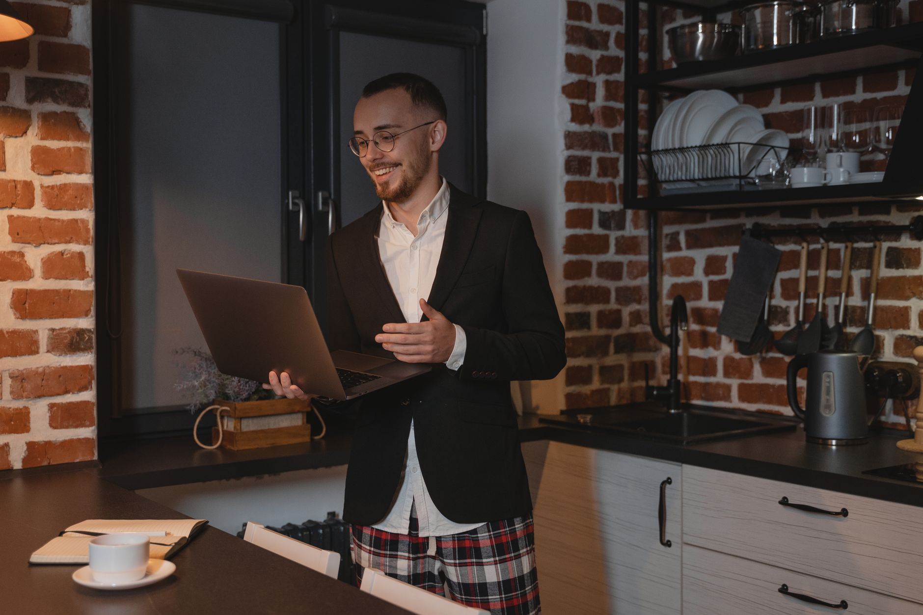 Lächelnder Mann in Jacke und karierten Shorts, der in einer Küche einen Laptop hält