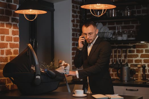Man in suit multitasks with baby in carrier while on a phone call in stylish kitchen.
