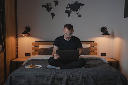 Man working on a laptop in a cozy bedroom setting, showcasing remote work lifestyle.