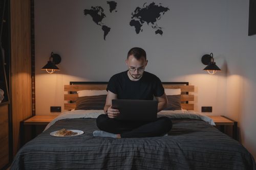 Free Man working on a laptop in a cozy bedroom setting, showcasing remote work lifestyle. Stock Photo