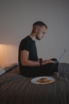 Side view of a young man working on a laptop while sitting on a bed, embodying remote work lifestyle.