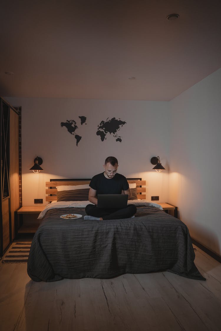 Man In Black Shirt Using A Computer Laptop Sitting On His Bed 