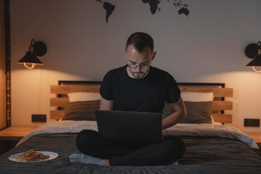 Man using laptop while sitting on bed with warm decor and wall map.