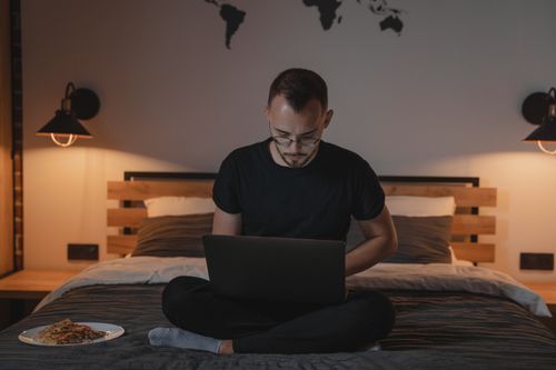 Free Man using laptop while sitting on bed with warm decor and wall map. Stock Photo