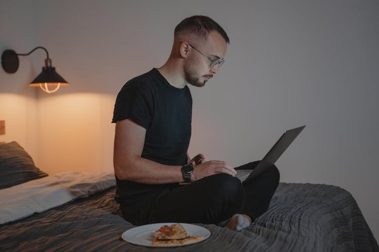 Man In Blue Crew Neck T-shirt Sitting On A Bed Using A Laptop Computer