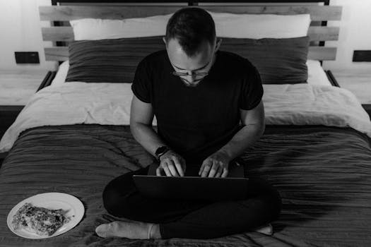 Man working on a laptop in a cozy bedroom, embracing remote work lifestyle.