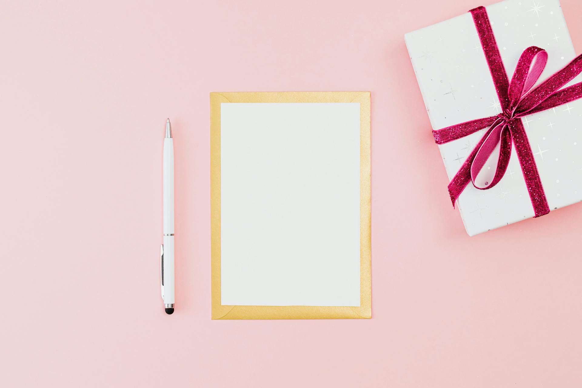 White gift box with magenta bow, gold-edged card, and pen on a soft pink background.