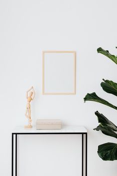 A minimalist home interior featuring a wooden figurine, frame, and potted plant against a white wall.