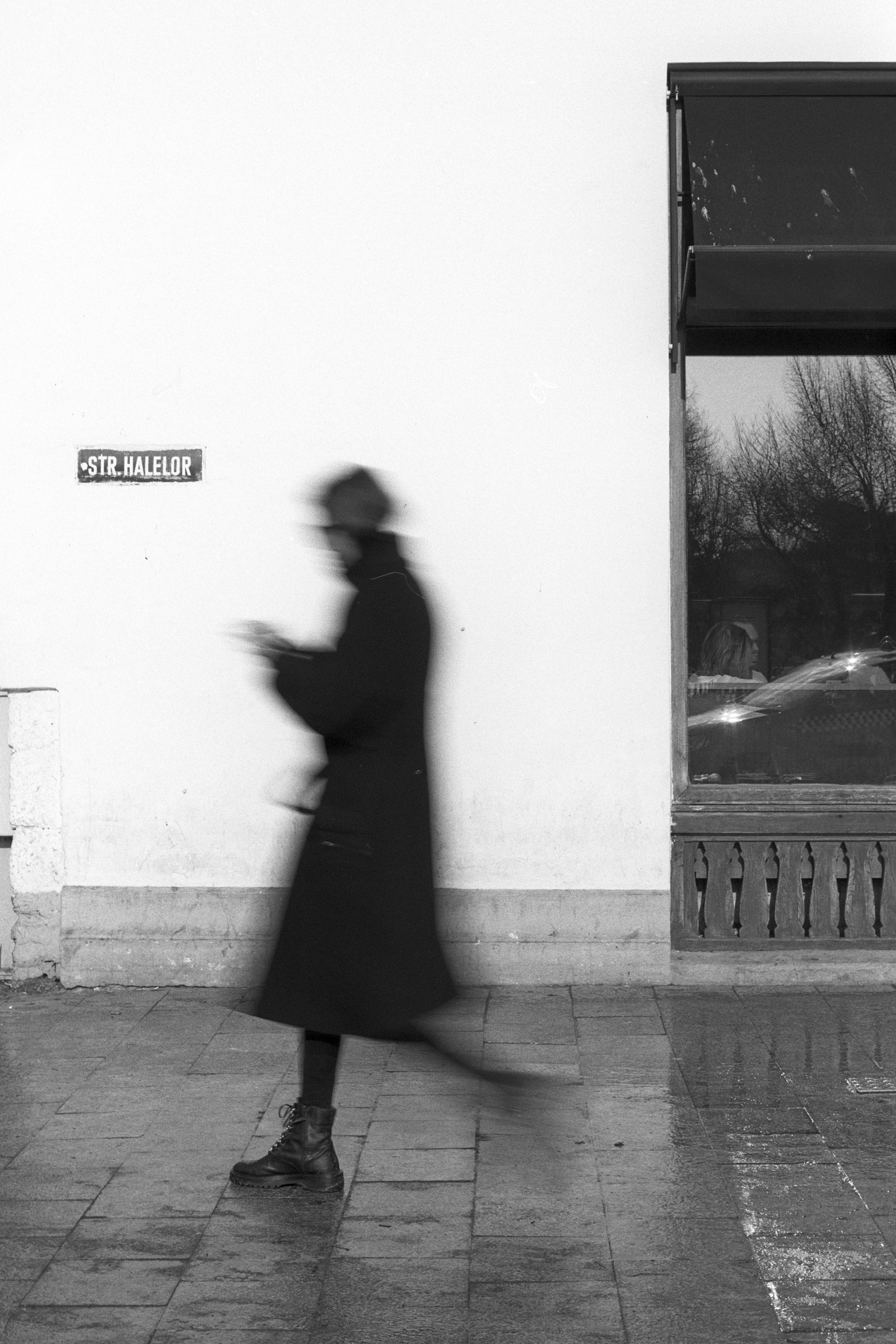 Black and white photo capturing a blurred figure walking on a sidewalk next to a window display.